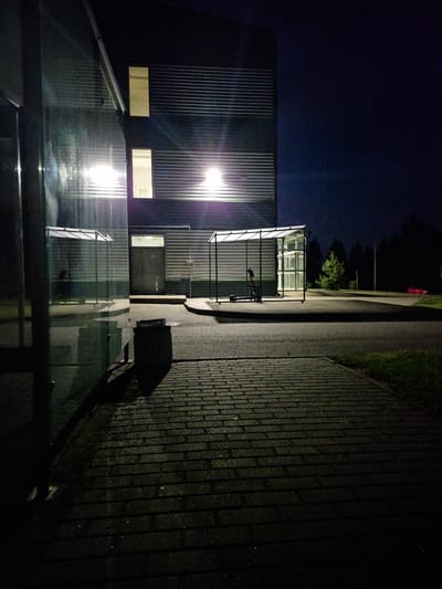 The image shows a dark, outdoor scene at night. In the foreground, a brick pathway extends towards the viewer, with shadows cast from unseen objects. To the left, a large glass structure with a corrugated metal roof reflects bright interior lights. Behind the glass structure, a modern building with horizontally ribbed dark siding is visible, featuring two illuminated windows. One window is a large, rectangular opening on the upper floor, and the other is a smaller, square window on the lower floor. A dark doorway is positioned between these windows. To the right of the building, a covered bus shelter with a transparent roof and white interior panels stands. Inside the shelter, a dark-colored scooter is parked. Further to the right, a red car is visible in the distance on a paved road. The background consists of dark trees and a clear night sky. The scene appears to be located in an urban or suburban area, potentially near a bus stop or public transportation hub, in Pabradė, Lithuania.