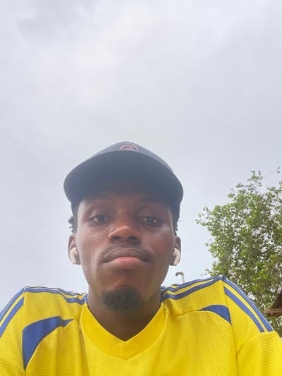 The image shows a close-up, waist-up selfie of a young Black man wearing a yellow and blue striped athletic shirt and a black baseball cap. He has white earbuds in both ears and appears to be looking directly at the camera. The background is mostly a cloudy sky with the top of a green tree visible on the right side. The man is in an outdoor setting, possibly in Osu, Nigeria, though the location is not explicitly confirmed in the image.