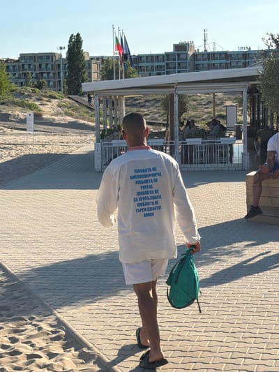 A man walks away from the camera on a paved walkway beside a sandy beach in Nesebar, Bulgaria. He is wearing a white shirt with blue text on the back and white shorts, and he carries a green backpack in his right hand. The text on his shirt reads: "ЛЮБОВТА НЕ Е СКОРОМЕРНИЧИ, ЛЮБОВТА НЕ РУПТАЕ, ЛЮБОВТА НЕ Е ПРЕВЪЗНАСЯ, ЛЮБОВТА НЕ ТЪРСИ СВОЕТО! АМИН" which translates to "Love is not hasty, love does not boast, love is not puffed up, love does not seek its own! Amen." In the background, there are buildings, trees, and people sitting at tables. The sky is clear and blue.