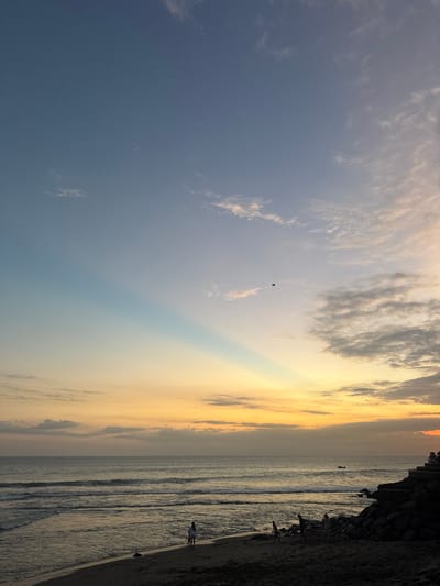 This is a vertical, eye-level, medium shot of a beach at sunset in Mengwi, Indonesia. The sky transitions from a deep blue at the top to a pale yellow and orange hue near the horizon. A wide beam of sunlight pierces the sky at an angle from the upper left. Clouds are scattered throughout the sky, with some appearing thin and wispy and others more dense and textured. A dark, diamond-shaped object, possibly a kite, is visible in the upper portion of the sky.

The ocean occupies the middle section of the image, with gentle waves breaking on the shore. The water appears dark and reflects the colors of the sky. The beach is in the foreground, with dark sand. A few scattered figures are present on the beach. On the left side, two small figures stand near the water's edge. Further to the right, three more figures are visible walking on the sand. On the far right, a rocky outcrop leads to a set of stairs where a small group of people are seated, silhouetted against the sky. The scene depicts a peaceful evening at the beach.