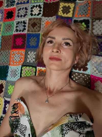 The image shows a woman with short, reddish-blonde hair and light skin. She is wearing a necklace with a small, diamond-encrusted crown pendant. Her left shoulder and upper chest are visible, and she appears to be wearing a garment with a colorful, abstract pattern. She has light makeup on, including mascara and lipstick. The background is a crocheted blanket with a patchwork of colorful squares. There is no discernible city or location information present in the image.