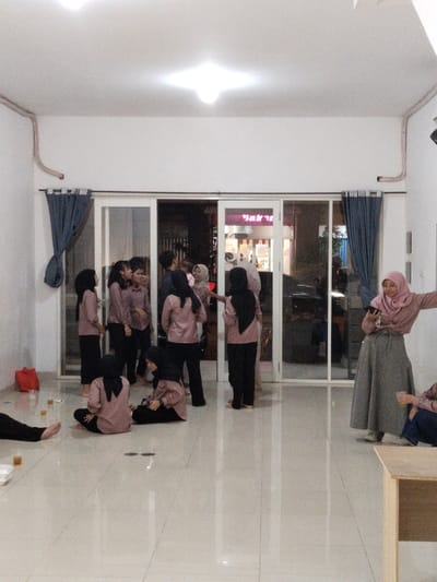 The image shows a group of 10 women in a room in a building located at coordinates -6.308, 107.282 in Telukjambe, Indonesia. The room has a white ceiling and white walls, with a tiled floor. There are large glass doors leading out to the street and a small shop with a pink sign that reads "Bakso" in the window. Two of the women are sitting on the floor near the glass doors, while the others are standing, talking to one another. One woman is using a mobile phone. There is a small table in the corner of the room, with a wooden tabletop and a single chair.