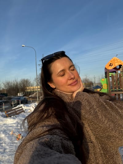 A female individual is depicted in a close-up, slightly low-angle shot, facing forward with her eyes closed and a serene expression, her chin resting on her right hand. She has long, dark hair, with black sunglasses perched on her head, and is wearing a brown textured outerwear piece over a light brown turtleneck. The right side of her face is illuminated by direct sunlight, casting a warm glow.
The scene is set outdoors in Akulovo (Gp Kubinka), Russia, on a clear, sunny day during winter. Snow covers the ground in the foreground and midground. To the left, a wooden park bench with light-colored slats is partially covered in snow. Beyond the bench, a paved area or street contains several parked vehicles, including a dark-colored sedan. White bollards or posts delineate the edge of the paved surface. Bare deciduous trees are visible in the background on the left.
To the right, a multi-colored children's playground structure, primarily orange, green, and purple, stands on the snow-covered ground. A street lamppost with a white fixture and a power line pylon are visible against the clear blue sky in the upper background. Overhead power lines traverse the sky. Distant structures and additional bare trees are present on the horizon. A small, orange and white toy is partially visible in the snow in the lower-left foreground.