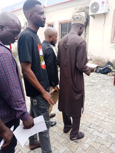 Four men stand outdoors on a paved area, possibly an Immigration Quarters in Nigeria. The man on the far left wears a plaid shirt and holds a white document. To his right, a man in a black t-shirt with a cartoon graphic is also holding paper. The third man, wearing a black suit jacket over a white shirt, has a shaved head and a beard. The man on the far right is dressed in a brown traditional garment and a patterned cap, and he is also holding papers.
The setting appears to be daytime, with neutral lighting suggesting it's not early morning or late evening. The ground is paved with light-colored interlocking bricks. In the background, there's a building with windows and an air conditioning unit mounted on the wall. Bags and shoes are visible on the ground behind the man in the brown garment. The men's expressions are neutral, and their postures suggest they might be engaged in a conversation or a briefing. There is no discernible text in the image besides the brand name "LG" on the air conditioning unit.