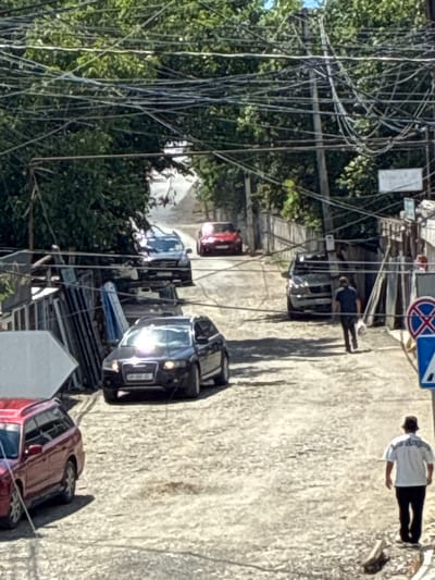 The image depicts a narrow, unpaved street in the city of Khashuri, Georgia, lined with buildings and overgrown with trees. A chaotic network of overhead wires dominates the upper portion of the scene. Several vehicles are present, including a dark-colored Audi sedan in the foreground, a red SUV to its left, a dark SUV further up the street, and a red sedan in the distance. Two men are visible walking on the street. One man, wearing a dark shirt and pants, is walking away from the camera towards a parked dark SUV, carrying a white plastic bag. Another man, wearing a white t-shirt with a graphic design and dark pants, is walking away from the camera towards the right side of the frame. Traffic signs are visible on the right side of the street. The scene suggests a residential or commercial area with a somewhat disheveled appearance.