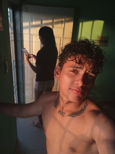 A low-angle, close-up selfie shows a young man with curly dark hair and a tattoo on his chest and neck. He is shirtless, and the light casts shadows across his face and body. In the background, a woman is silhouetted as she holds a phone and looks down at it. She is wearing a t-shirt and patterned shorts. The background appears to be an interior doorway with a white door, and a green wall is visible on the right. The overall lighting suggests it may be late afternoon or early evening, with sunlight streaming in from an unseen source. The location is identified as La Guardia, Venezuela.