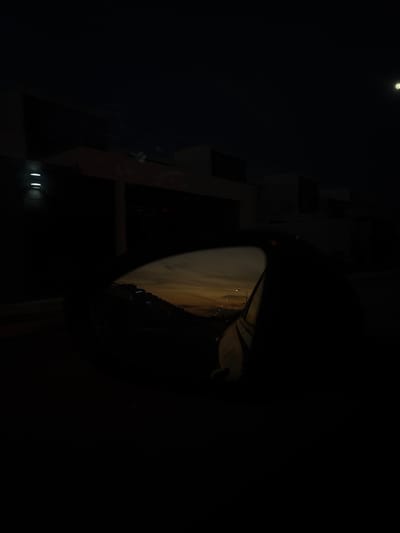 A very dark, low-lit image shows a convex mirror reflecting a cityscape at dusk or dawn. The mirror is predominantly black, obscuring the details of its surroundings. A tall building's structure is faintly visible to the left of the mirror, with dim lights illuminating its lower portion. A single light source, possibly the moon, appears in the upper left corner of the frame.
