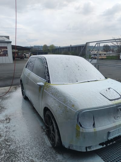 The image shows a car covered in soap suds, likely undergoing a car wash, at an outdoor location in Bindlach, Germany. The car's rear is visible, along with a tire. In the background, there is fencing, a building with signage in German, and trees. The sky is overcast. A red hose is visible on the ground.