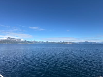 This media file shows a vast expanse of deep blue ocean under a clear blue sky dotted with scattered white clouds. The water is slightly rippled, suggesting a gentle breeze. In the distance, a majestic mountain range forms a dramatic backdrop. The mountains are rugged and appear to be covered in patches of vegetation and possibly snow at their peaks. Some of the mountains have sharp, pointed summits, while others are more rounded.
The scene is bathed in bright sunlight, indicating it is daytime. The clear sky and calm water suggest pleasant weather conditions. The overall impression is one of natural beauty and tranquility. The perspective is from a body of water looking towards land, possibly from a boat or the shore. There are no visible people or man-made objects other than what might be implied by the viewpoint.
The location context provided, Hierenjárgga - Korsnes, Norway, places this scene in a region known for its stunning fjords and mountainous landscapes. This context aligns with the visual elements of the image, characterized by the sea meeting the dramatic coastline of Norway.