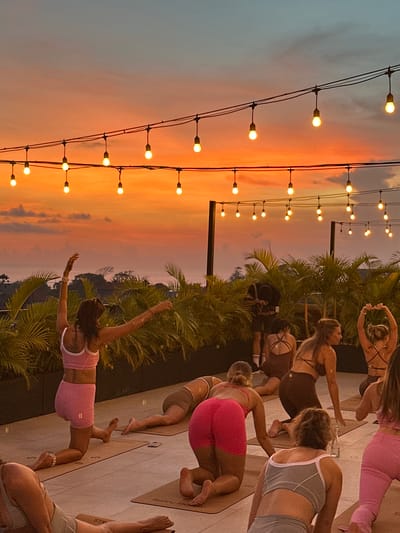 The image shows a group of people participating in a yoga session outdoors in Tibubeneng, Indonesia. The yoga participants are positioned on yoga mats on a tiled patio. Most are women. They are wearing yoga attire. In the background are palm trees and a sunset. String lights are visible in the foreground. A photographer appears to be present.