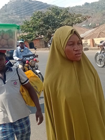 A woman wearing a yellow hijab is featured in the upper portion of the image, a motorbike with two men riding in the lower portion of the image. The background includes buildings, trees, a large hill, and overhead power lines. The men on the motorbike appear to be driving on a road. The light and perspective of the photo suggest it was taken in daylight, outside.