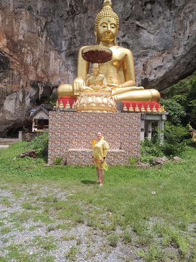 The image depicts a large, golden Buddha statue situated in front of a rocky cliff face in Thap Put, Thailand. A smaller Buddha figure sits beneath the larger one. A woman wearing a yellow raincoat stands in front of a tiled wall located in front of the Buddha statue. The ground consists of grass and gravel. A small building is visible to the left of the statue. The weather appears overcast. Green foliage surrounds the area.