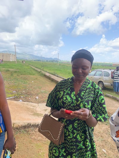 In Gwafan, Nigeria, a young woman in a black and green patterned dress stands outdoors, holding a red object, possibly a lip balm or a small cosmetic. She wears a black head covering and a gold watch. A brown purse with pearl embellishments is slung over her shoulder. In the background, a silver SUV is parked, and a man in a striped shirt and blue jeans stands beside it. The setting appears to be a semi-urban area with green fields, a dirt path, and distant hills under a cloudy sky.
