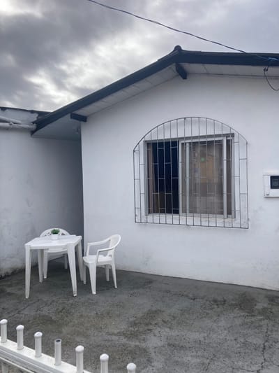This media file captures an outdoor scene, likely a patio or side yard, next to a white house in Florianópolis, Brazil. The image is composed primarily of a white stucco wall, a dark roofline, a concrete ground, and an overcast sky.
A prominent feature is a rectangular window set into the white wall, protected by an ornate metal security grate. The grate is silver-colored, formed from a grid of horizontal and vertical bars, with its top and left side featuring a large curve. Above the window, a small, white rectangular box, possibly an electrical fixture or light, is mounted on the wall.
Below the window, on the concrete ground, is a small outdoor dining or seating area. It consists of a white plastic table with a small green plant in the center, flanked by two white plastic chairs. One chair is pulled up to the table, while the other is positioned slightly away.
To the far left, a dark roofline with a gutter system runs across the frame, with a blue electrical cable visible running along it and down the side of the building. The sky above this roofline is heavily overcast, filled with thick gray clouds, suggesting a cloudy day or impending rain.
The concrete ground is light gray, showing signs of wear, cracks, and appears damp in places, possibly from recent rain or moisture. To the far right, a segment of a white railing with several decorative, cylindrical posts is visible. No people are present, and no discernible text is visible in the image.