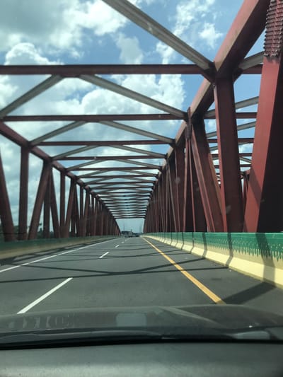The image depicts a view from a vehicle moving along a bridge. The bridge's structure is made of red metal beams arranged in a geometric pattern against a cloudy sky. The road surface has white lane markings and a yellow dividing line. Part of the vehicle's interior is visible on the right side of the frame. The location is implied to be Gohor Lama, Indonesia, based on the prompt.