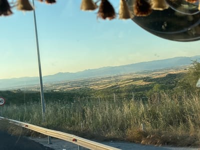The image shows a rural landscape in Avra, Greece, viewed from inside a vehicle. In the foreground, a metal guardrail runs along the side of a road, with tall, dry grass and wildflowers growing on the embankment. A grey pole, likely a streetlamp or utility pole, stands tall on the right side of the road. A circular speed limit sign with the number "90" is visible on the left. The background reveals a vast valley with rolling hills and agricultural fields, segmented by trees and vegetation. In the distance, mountains form a hazy silhouette against a clear, light blue sky. The scene is illuminated by natural sunlight, suggesting it was taken during the daytime. The overall impression is one of a serene, open countryside.