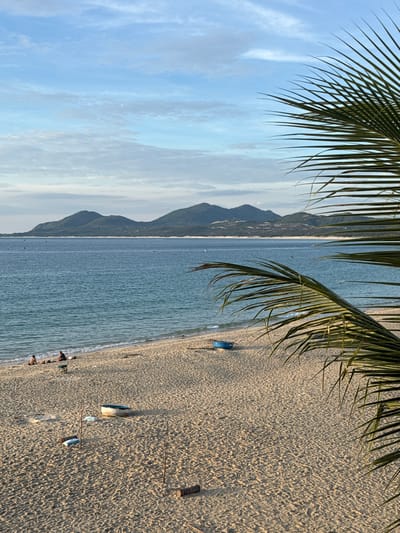 This is an eye-level, outdoor, vertical shot of a sandy beach with the ocean and mountains in the background. The scene is captured in Song Cau, Vietnam, likely during the late afternoon or early evening due to the warm lighting.
In the foreground, a palm frond partially obscures the view from the right. The sandy beach is textured with small ripples, indicating recent tides or wind. Several objects are scattered across the sand: a blue, oval-shaped boat rests on its side near the center-left, and closer to the bottom right, two rough, wooden poles are stuck into the sand, connected by a taut rope, forming a makeshift net or boundary. Two cylindrical objects, appearing to be rolled mats or logs, lie on the sand. One is positioned near the base of the poles, and another is further left, between the boat and the poles. Two white, flat objects, possibly towels or cushions, are placed on the sand beneath the rope.
Further back, near the water's edge, a small group of people are visible. Two individuals are seated on the sand, facing the ocean, with another person sitting between them. A small green stool or table is positioned in front of them. A few other figures are dotted along the shoreline.
The ocean is a calm, turquoise blue, with gentle waves lapping at the shore. A single, small blue boat is visible in the distance on the water. Beyond the ocean, a line of dark green, rolling mountains forms the horizon, their slopes dotted with patches of lighter vegetation and some structures. The sky above is a soft blue, with scattered, wispy white clouds that are illuminated by the setting sun, creating a serene and peaceful atmosphere.