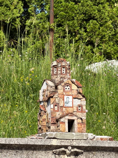 A miniature, rough-hewn stone church, possibly a roadside shrine, stands amidst tall green grass and wildflowers. The church is constructed from irregularly shaped stones and bricks, with a pitched roof. A small framed image of a woman holding a child is affixed to the facade. The shrine sits on a grey, weathered stone base, which is part of a larger stone structure in the foreground. In the background, dense green trees form a backdrop, with a rusty metal pole visible near the center. The scene appears to be outdoors, likely in a rural or semi-rural setting, possibly in Barziya, Bulgaria. The lighting suggests a sunny day.