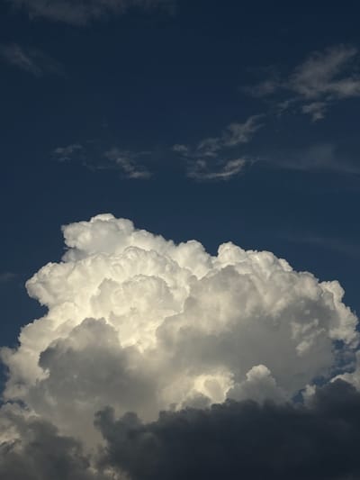This is a photograph of a dark blue sky filled with dramatic clouds. On the right side of the frame, a large, bright white cumulus cloud dominates, its textured surface illuminated by sunlight. The cloud has a voluminous and somewhat chaotic appearance, with billows and folds creating a sense of depth. To the right of this bright cloud, a darker, more ominous cloud bank is visible, appearing as a deep gray mass that obscures the sky. Thin, wispy white clouds are scattered across the left side of the dark blue sky, appearing more distant and less substantial than the main cloud formation. The contrast between the bright sunlight on the cumulus cloud and the darkness of the storm clouds suggests dynamic weather conditions. The image has a vertical orientation, emphasizing the height and grandeur of the sky. There is no visible text. The overall mood is dramatic and awe-inspiring, evoking the power of nature. Given the location context of Novy, Russia, this could be a scene captured during a summer storm. The time of day is likely late afternoon or early evening, as indicated by the strong contrast and directional lighting on the clouds.