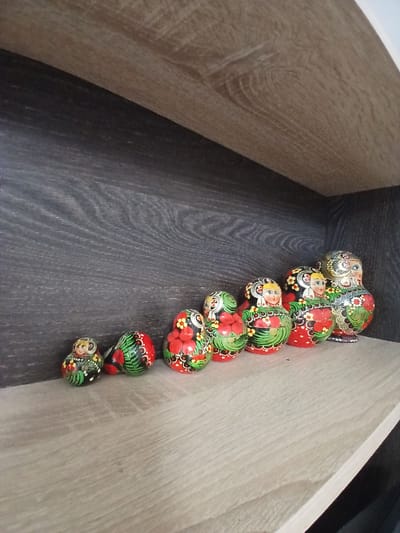 This is a close-up, eye-level, indoor shot of a collection of Matryoshka dolls arranged on a wooden shelf. The dolls are nested and vary in size, with the smallest on the left and the largest on the right. They are painted with traditional Russian folk art designs, featuring bright colors like red, green, yellow, and black, with floral patterns and stylized faces. The shelf itself is made of light-colored wood with a visible grain, and it is set against a darker, wood-grained background, possibly another shelf or a wall. The lighting is neutral, creating soft shadows. There is no indication of the specific location in Maslova Pristan', Russia, beyond the implicit context that Matryoshka dolls are a Russian cultural item.