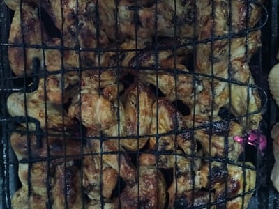 A close-up, overhead view shows a grill basket filled with numerous pieces of grilled chicken wings. The chicken is a golden brown color with some darker, charred areas, indicating it is well-cooked. The grill grate is visible, with black metal bars crisscrossing over the food. In the background, a faint purple glow is visible, suggesting the presence of coals or a heat source beneath the grill. The image captures a food preparation scene, likely in an outdoor setting given the grilling apparatus. There are no people visible in the image. The scene is devoid of any specific landmarks or identifying features that would place it in the city of Niš, Serbia.