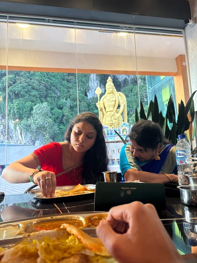 A photo shows two women and a hand sitting at a table with a view of a park and a gold statue. The women are eating off of trays of food. The woman closest to the camera is wearing a red top and has dark hair. The woman further back has gray hair and is wearing a blue top. The hand on the right is reaching for food. The scene is in the city of Selayang Municipal Council, Malaysia.