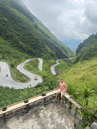 A man stands on a paved overlook next to a wooden railing, gazing toward the camera. He wears a pink t-shirt, khaki shorts, and a baseball cap. Below, a winding road with several hairpin turns traverses a steep, lush green hillside. A white car and a motorcycle are visible on the road. The sky is overcast. The location is Phố Bảng Commune, Vietnam.