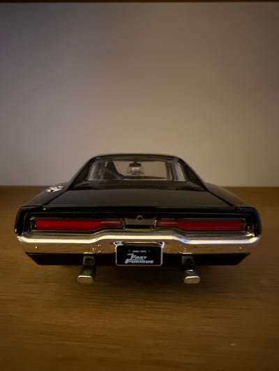 The image shows the rear view of a black toy car, likely a model of a Dodge Charger from the "Fast and Furious" franchise, resting on a wooden surface. The car features a prominent red taillight bar and chrome bumper. A license plate with "THE FAST AND FURIOUS" printed on it is visible. The background is a plain, dimly lit wall. There is no indication of the city of Bruges, Belgium, in the image.
