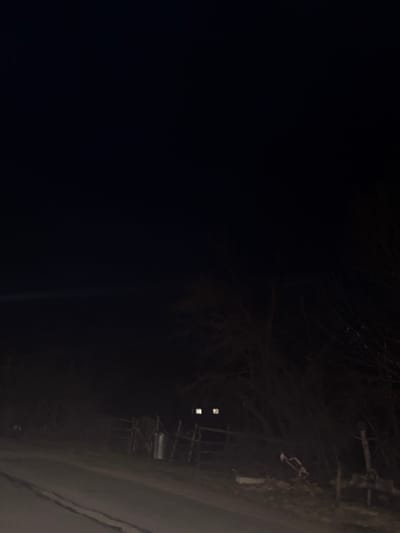 The image depicts a static nighttime scene in Iamna, Bulgaria. A paved road occupies the lower portion of the frame, extending into the dark background with visible faint markings. To the left of the road, a rustic wooden fence, composed of vertical posts and horizontal rails, is present. Adjacent to the fence, a metallic, cylindrical object, possibly a gas canister or bin, stands upright. Beyond the fence and further into the midground, a building is partially visible through bare deciduous trees. This building features two brightly illuminated, rectangular windows emitting a distinct white light. The right side of the frame is dominated by a dense cluster of bare trees and shrubs. The upper half of the image is completely dark, indicating a nocturnal environment. No individuals are visible. A small, light-colored, indistinct object rests near the base of the trees on the far right side of the road.