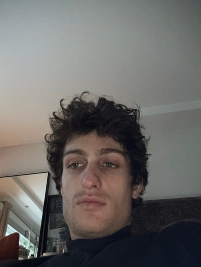A male individual is depicted in a close-up, slightly upward-angled view within an indoor setting. He has dark, curly hair and light-colored eyes, which are directed downward and to the viewer's left. His nose has a slight reddish tint at the tip, and he exhibits subtle stubble on his face. He is wearing a dark, high-necked garment, likely a turtleneck. The upper portion of the image shows a plain, light-colored ceiling. To the left of the subject, a large, dark-framed mirror reflects a window with vertical elements, and a hint of an orange-red soft furnishing is visible. The right side of the background shows a dark, textured surface, possibly part of a furniture piece. The lighting is even, suggesting ambient indoor illumination. The specific location within Vincennes, France, cannot be determined from the visual information presented in the image.