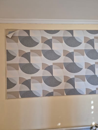 The image depicts a section of wallpaper with a geometric pattern of circles and rectangles in shades of gray, beige, and off-white. The wallpaper is partially peeled away from the wall at the top left corner. The wall beneath the wallpaper appears to be a light yellow color. Above the wallpaper, there is white crown molding. The scene is indoors, likely a room in a residential or commercial building. The lighting suggests it is daytime, with some shadows cast on the yellow wall from an unseen light source. There are no people or animals visible in the image. The location of Bereke, Kazakhstan is not discernible from the visual information provided in the image.