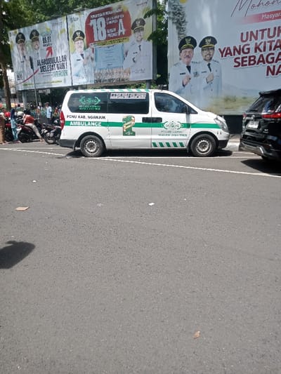 The image depicts a daytime street scene in Nganjuk Regency, East Java, Indonesia. In the foreground, a dry asphalt road with a visible white line features scattered small debris and a dark, indistinct shadow on the left. A white van, clearly marked as an "AMBULANCE" for "PCNU KAB. NGANJUK" and "NU CARE-LAZISNU NGANJUK JAWA TIMUR LUXURY CLASS" with green decals and the Nahdlatul Ulama (NU) logo, is parked on the right side of the road. To the immediate right and slightly behind the ambulance, the rear bumper and wheel of a black SUV are partially visible. On the far left, a small group of individuals, some wearing headscarves, are standing near multiple parked motorcycles. In the background, three large billboards are prominent, each displaying images of uniformed men, likely local officials, alongside text in Indonesian. Green tree foliage is visible above and between the billboards.