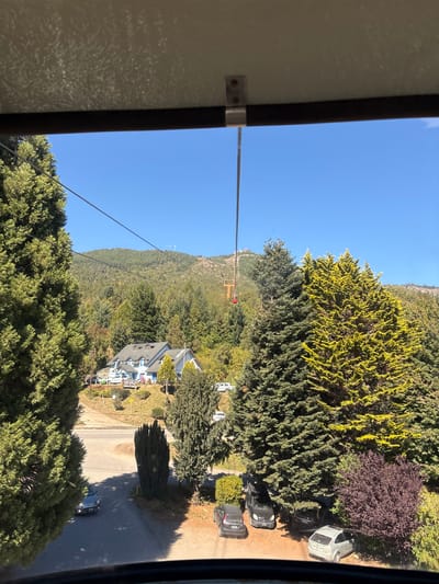 The image depicts an elevated view of a mountainous, residential landscape in San Carlos de Bariloche, Argentina, captured from inside a gondola cabin. The sky is clear and brilliant blue, dominating the upper frame. Below the sky, a verdant mountain range covered densely with coniferous trees, some exhibiting a yellowish-green hue, extends into the background.

A prominent light-blue, multi-story building with white trim, a dark grey roof, and multiple dormer windows is situated centrally on a gently sloping hill. Surrounding the building are various trees, shrubs, and a winding paved road. Multiple vehicles are visible; a silver hatchback is parked on the paved road in the lower-left, and several others are parked on unpaved surfaces: a black van, a dark sedan, another dark vehicle, and a light-colored SUV.

A gondola cable diagonally traverses the scene from the lower-left towards the upper-right. A single red gondola cabin is visible mid-air in the mid-ground, ascending towards a distant mountain peak where a chairlift tower and other smaller structures are discernible. No individuals are explicitly present in the frame, but the infrastructure and vehicles indicate human presence and activity within the environment.