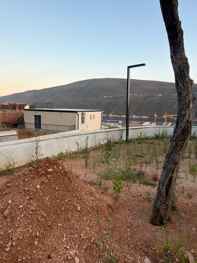 The image, taken in the city of Đenovići, Montenegro, depicts an outdoor scene under a clear sky. In the foreground, a pile of reddish-brown soil and rocks is visible, with sparse green vegetation growing around it. To the right, the textured bark of a tree trunk is prominent. A modern, light-colored building with dark-framed windows and a railing system is situated on a raised concrete platform behind a low, white wall. A dark, rectangular lamppost stands to the left of the tree. In the background, a body of water with several construction cranes and boats is visible, beyond which a tree-covered hill slopes upwards. The sky transitions from a pale yellow near the horizon to a light blue overhead. The scene appears to be a construction or development area.