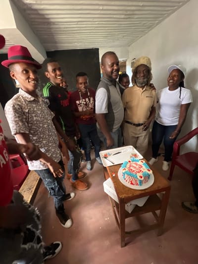 The image shows a group of seven people gathered in what appears to be a residential or commercial building in Githurai, Kenya (coordinates -1.205, 36.904). The scene depicts a birthday celebration as evidenced by a cake on a table in the center of the frame with blue and red frosting and the words "Happy Birthday" written on it. The individuals, who are all dressed casually, are interacting with one another, with a few of them smiling. The person in the front is wearing a black and white patterned shirt, blue jeans, and dark sneakers. They are holding a red chair in their right hand. To the left of them are two men in similar clothing, one wearing a hat and the other with a brown and white shirt. In the background, three other individuals are standing near a table with a cake on it. One is wearing a white shirt, and the two others have their backs to the camera. The environment consists of a white-walled room with a wooden floor and wooden furniture. The room appears to be sparsely decorated. The overall scene is one of celebration and camaraderie.