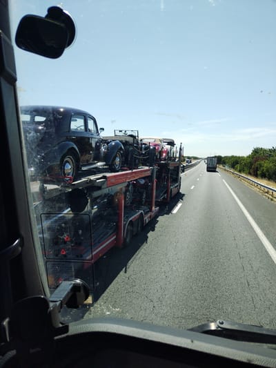 A car carrier truck transports several vintage cars down a highway under a clear blue sky. The perspective is from the driver's seat of another vehicle, looking forward and slightly to the right. The car carrier is loaded with at least three cars: a black sedan on the upper deck, and a red convertible and a white car partially visible on the lower deck. The highway appears to be in a rural area, with trees lining the right side of the road. A white truck is visible in the distance on the highway. The scene is consistent with traffic on the A20 autoroute near Déols, France, a common route for vehicle transport.