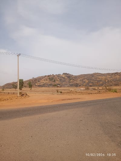 The image depicts an outdoor, semi-arid landscape located in Barakin Columbai, Nigeria, captured at 10:35 on October 2, 2026. The foreground features a paved asphalt road, dark grey with visible aggregate, extending across the lower half of the frame. To the immediate right of the road is a wide, flat expanse of reddish-brown dirt. A light-colored utility pole stands vertically on the left side of this dirt area, supporting four parallel overhead power lines that stretch horizontally across the mid-frame. Behind the open dirt plain, a low, rocky mountain range or extensive hillside rises, characterized by exposed rock formations and sparse, dry vegetation. Several small, sparsely leaved trees are scattered within the midground plain and near the base of the hillside. Minor concrete foundations are visible in the dirt area in the mid-distance. The sky above is overcast with diffuse light. No individuals, vehicles, or active subjects are present.