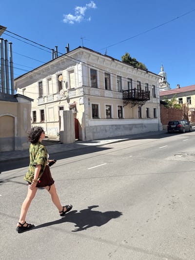 In Yelets, Russia, under a bright blue sky with a few wispy clouds, a person with curly dark hair walks across a street. They are wearing sunglasses, a green shirt with a tropical print, and a brown skirt. Black sandals are on their feet, and they are holding a dark-colored smartphone in their right hand. The person casts a long shadow on the asphalt road, which has white lane markings.
Behind the person is a street sidewalk and a light-colored, multi-story building with ornate architectural details. The building has several windows, some with brown frames. A black wrought-iron balcony juts out from the upper floor. Adjacent to this building is another structure, partially visible, with tall, grey, cylindrical chimney-like objects protruding from its roof. Further in the background, other buildings are visible, including one with a prominent white dome and spire, suggesting it might be a church or cathedral. A dark-colored SUV is parked on the right side of the street, near a brick wall. The overall scene depicts a sunny day in an urban environment.