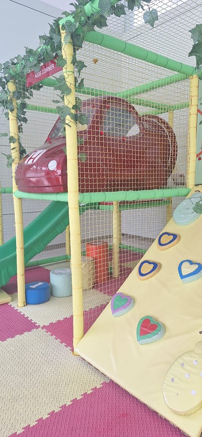 The image depicts a colorful indoor playground area designed for children, likely located in a commercial establishment or recreational facility in Babakan Madang, Indonesia. The playground features a large, red car-shaped structure serving as a playhouse or tunnel, enclosed within a yellow and green netting structure adorned with artificial green leaves. Adjacent to the car structure is a bright green slide. The floor is covered with interlocking foam mats in pink, white, and yellow patterns, providing a soft landing surface. Several cylindrical poufs in blue and light blue, labeled with numbers, are visible near the slide. The foreground shows a yellow padded ramp, part of a climbing structure, with several colorful heart-shaped handholds and footrests attached. The overall environment is bright and playful, suggesting a safe and engaging space for children's activities.
