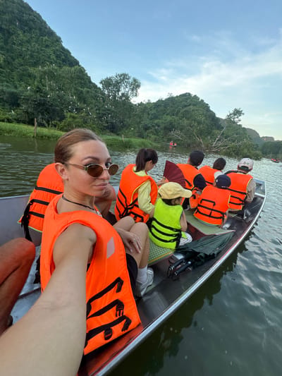 In Yên Sơn Ward, Vietnam, a group of people are on a boat tour on a river, surrounded by lush green hills and trees. The water is calm and reflects the sky. Several individuals are wearing orange life vests, suggesting a tourist activity. In the foreground, a woman is taking a selfie, looking at the camera with sunglasses on. She is wearing an orange life vest. Other people in the boat are facing away from the camera, looking at the scenery. Some are children wearing bright green vests. There are birds visible in the trees on the hillsides. The overall scene depicts a leisurely boat excursion in a natural setting.