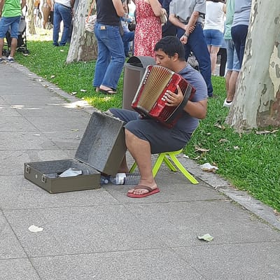 A man is playing an accordion while sitting on a bright green plastic stool on a paved walkway. He is wearing a grey t-shirt and dark grey shorts, and red flip-flops. The accordion is red and has a black strap. Next to him is an open, olive-green suitcase containing a white plastic bag. Two plastic water bottles are on the ground near the suitcase. The man is outdoors, with green grass and trees in the background. The walkway appears to be in a park or public space. The scene suggests a casual outdoor setting, possibly a street performance or a moment of leisure. The overall impression is one of everyday urban life.