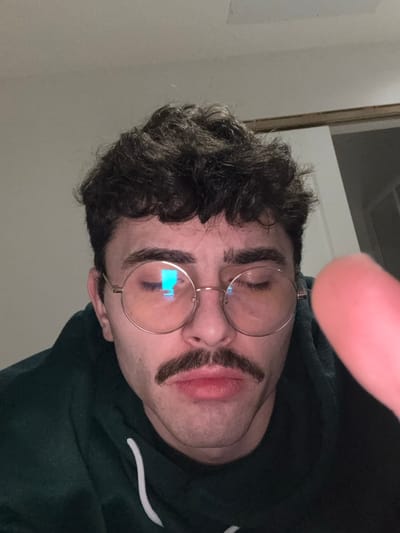 A person with curly dark hair is wearing round eyeglasses. Two dark, arched lines mimic a mustache and cover the lips. The individual wears a dark green hooded sweatshirt with a white drawstring visible. A blurry finger is in the top right corner of the frame. The background is a light-colored wall and part of a door frame is visible. The image is oriented sideways.