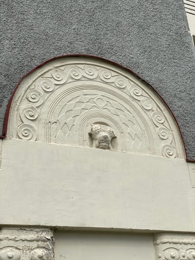 This is a close-up, eye-level, outdoor photograph of architectural ornamentation on a building in Sortavala, Russia. The subject is a carved relief in a light color, possibly stucco or plaster, set against a textured, gray exterior wall. The relief is semicircular and features a central depiction of an animal's head, possibly a lion or dog, with an open mouth and prominent teeth. Surrounding the head are concentric semi-circular bands with decorative patterns, including swirling motifs and geometric shapes resembling leaves or feathers. The relief is partially weathered, with some cracking and peeling of the surface. A thin, dark red band borders the top edge of the relief. Below the relief, a smooth, light-colored horizontal surface extends across the frame, also showing signs of cracking. The bottom of the image reveals further decorative elements with a similar worn appearance. The environment appears to be overcast, judging by the diffuse lighting. There are no people or animals present in the image.