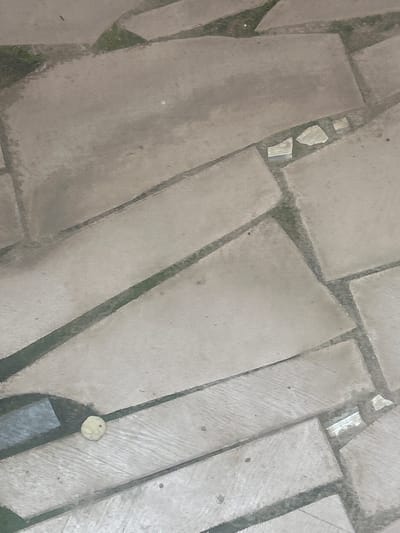 This is an aerial, oblique photograph of a stone-paved walkway. The paving stones are irregularly shaped and of varying sizes, laid with dark grout or soil between them. Some of the paving stones have a rough, textured surface, while others appear smoother. There are patches of green vegetation growing in the gaps between some of the stones. In the upper left quadrant, there are a few smaller, lighter-colored stones. In the lower right quadrant, there is a small, round, light-colored object, possibly a pebble or a piece of discarded material, next to a darker, rectangular shape which may be a shadow or a wet patch. The lighting suggests it is daytime, and the overall impression is of an outdoor area, possibly a pathway in a garden or a public space. No people are visible, and no text is present in the image. The setting appears to be in Ogbomosho, Nigeria, as per the location context.