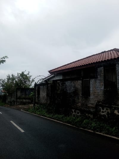 A weathered brick house sits alongside a wet asphalt road marked with a white line. The house, topped with red roof tiles, shows signs of age with stains and vegetation clinging to its walls. A wall extends from the house, also aged and covered in moss and plants. Overcast skies dominate the scene, contributing to the somber mood of the image.