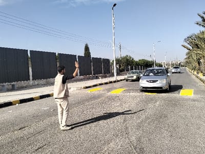 The image depicts a daytime scene on an asphalt road in the city of Homs, Syria, under a clear blue sky. A male individual, positioned in the foreground, stands on the road facing right, wearing a light-colored vest with a green logo on the chest over a dark shirt, and light-colored trousers. His left arm is raised with the palm open and forward, performing a stopping or halting gesture. His shadow is cast forward onto the road surface.
The road features two prominent yellow speed bumps in the midground. A silver sedan is positioned directly in front of these speed bumps, facing away from the camera. Further down the road, an older light-colored sedan is visible in the left lane, and a white SUV or sedan is in the right lane. The road is bordered on both sides by curbs painted with yellow and black stripes.
To the left of the road, a dark gray corrugated metal fence is visible, behind which a tall, slender coniferous tree and other foliage are present. Overhead power lines span the sky above the fence. To the right, a median strip contains multiple palm trees and other greenery. Several modern streetlights with curved designs extend into the distance, where urban buildings are faintly discernible against the horizon.