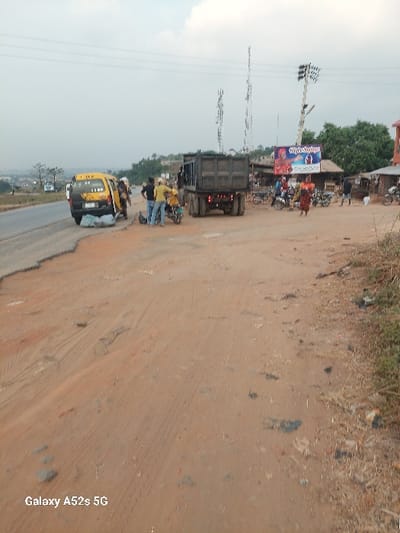 The image depicts a roadside commercial area in Awkuzu, Nigeria, under an overcast sky. The foreground consists of reddish-brown unpaved dirt exhibiting numerous tire tracks, transitioning to a worn asphalt road on the left. A yellow and white commercial passenger van, bearing "AWK" on its side and a white package secured to its rear, is parked on the shoulder of the paved road. Immediately behind it, a dark-colored dump truck is parked partially on the dirt shoulder and partially on the road.
Between these two vehicles, several individuals are gathered, including one man in a yellow shirt and blue jeans, standing near a yellow motorcycle. Approximately ten to fifteen people are visible across the scene. To the right, a cluster of rudimentary market stalls flanks the dirt area. Numerous motorcycles, commonly known as Okadas, are parked throughout this market section. One person in an orange top and patterned skirt walks towards the foreground from this area.
Prominently displayed on a wooden pole is a large billboard featuring a blue background and product advertisements. Two tall, metal telecommunication towers are positioned behind the dump truck and market stalls. A utility pole with multiple cross-arms and lights stands near the billboard. Power lines are visible stretching across the upper portion of the frame. The background shows distant hills with sparse vegetation. Individuals are observed standing, conversing, and moving within this bustling roadside environment.