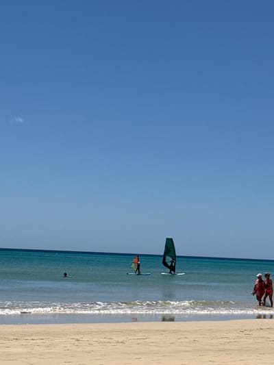The image depicts a beach scene in Pájara, Spain. The sky is clear blue, contrasting with the tan sand of the beach on the right side of the image. In the ocean, there are two windsurfers with colorful sails. In the distance, close to the beach, a group of people are standing near the water. The water itself is a light blue, with visible waves approaching the shore. A solitary figure is seen floating further out in the water.