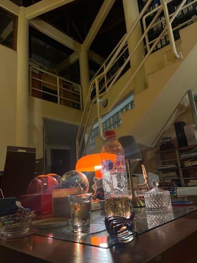 The image captures a cluttered table inside a building, likely a residence or office. On the table, a variety of objects are visible, including a plastic water bottle, a disco ball, various glasses and containers, books, and decorative items. The background shows architectural details such as structural beams, railings, and windows. Based on the limited information, the location is unidentifiable to be Santiago de Surco, Peru.