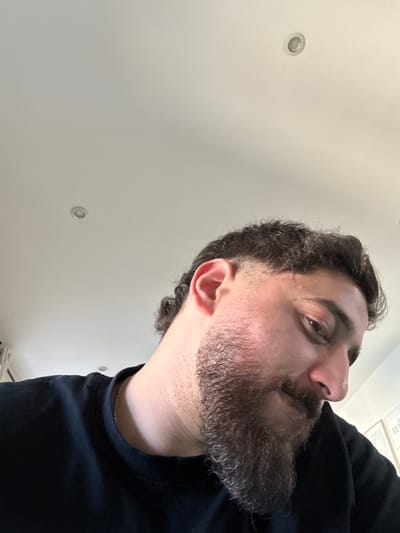 The image depicts a male individual in Lingolsheim, France, captured from a low-angle perspective. The subject has dark, curly hair with a faded cut on the sides and longer, curly hair at the back. A dark, full beard covers his lower face. He is wearing a black crew-neck t-shirt, and a thin, light-colored chain is visible around his neck. The individual's head is tilted downwards and to the right, with his gaze directed towards the lower right of the frame. A faint smile is visible on his face. The background consists of a plain, light-colored ceiling, featuring two circular recessed lighting fixtures. A blurred, partially visible white object is present in the bottom right corner of the frame.